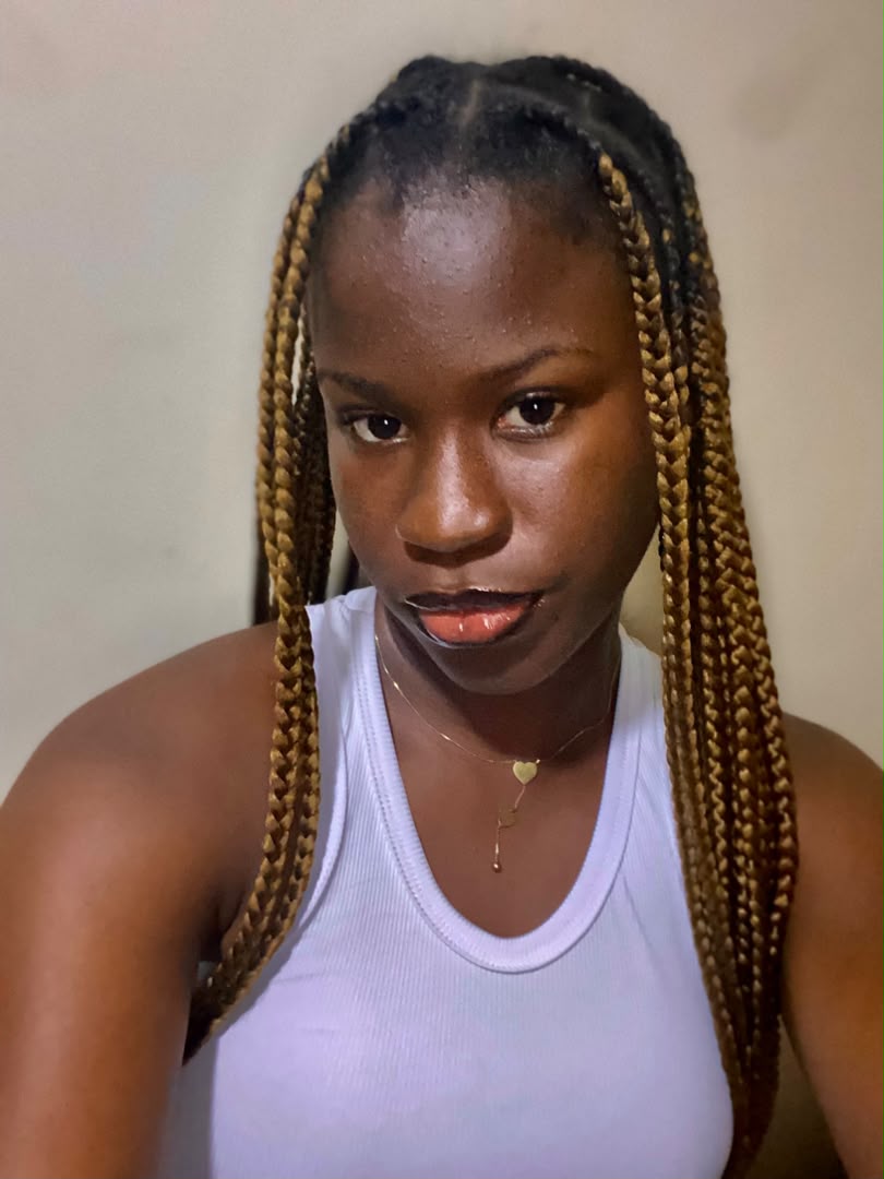 Divine in a white tank top with long honey-blonde box braids, looking straight into the camera.