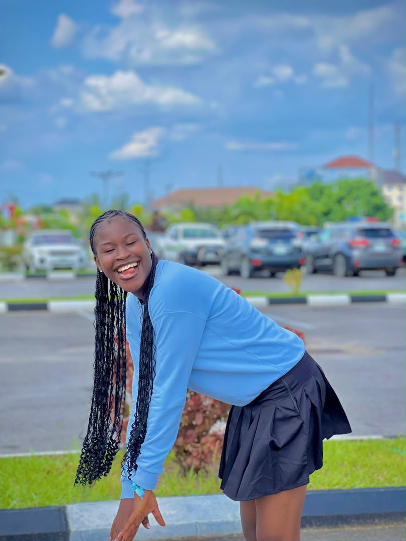 Divine in a blue sweatshirt and black skirt, bent over laughing under a wide cloudy sky.