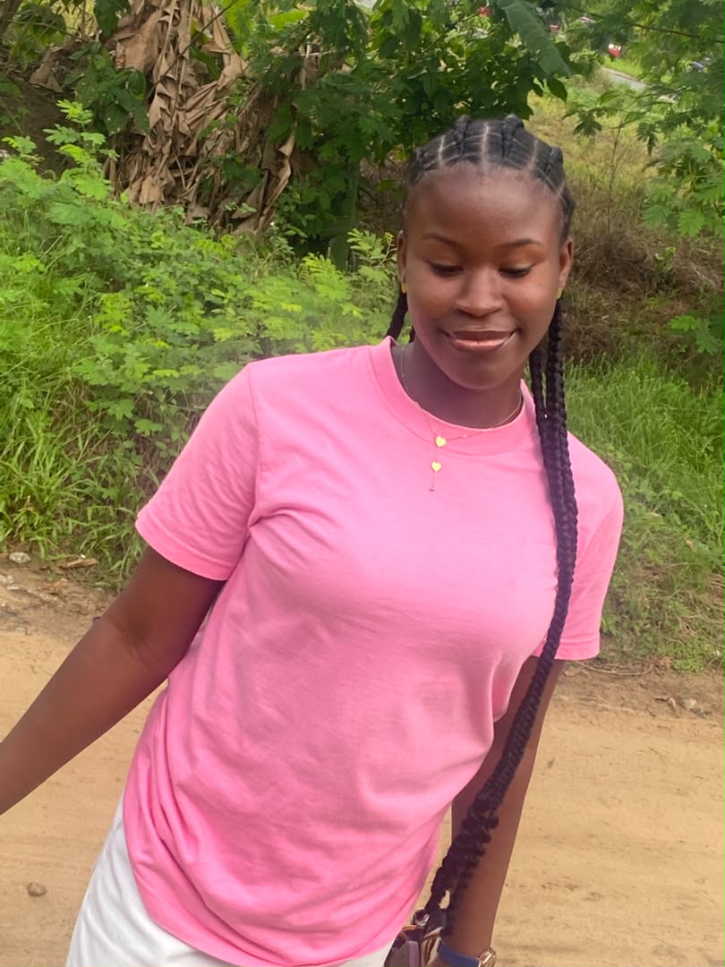 Divine in a pink t-shirt, looking down and smiling, greenery behind her.