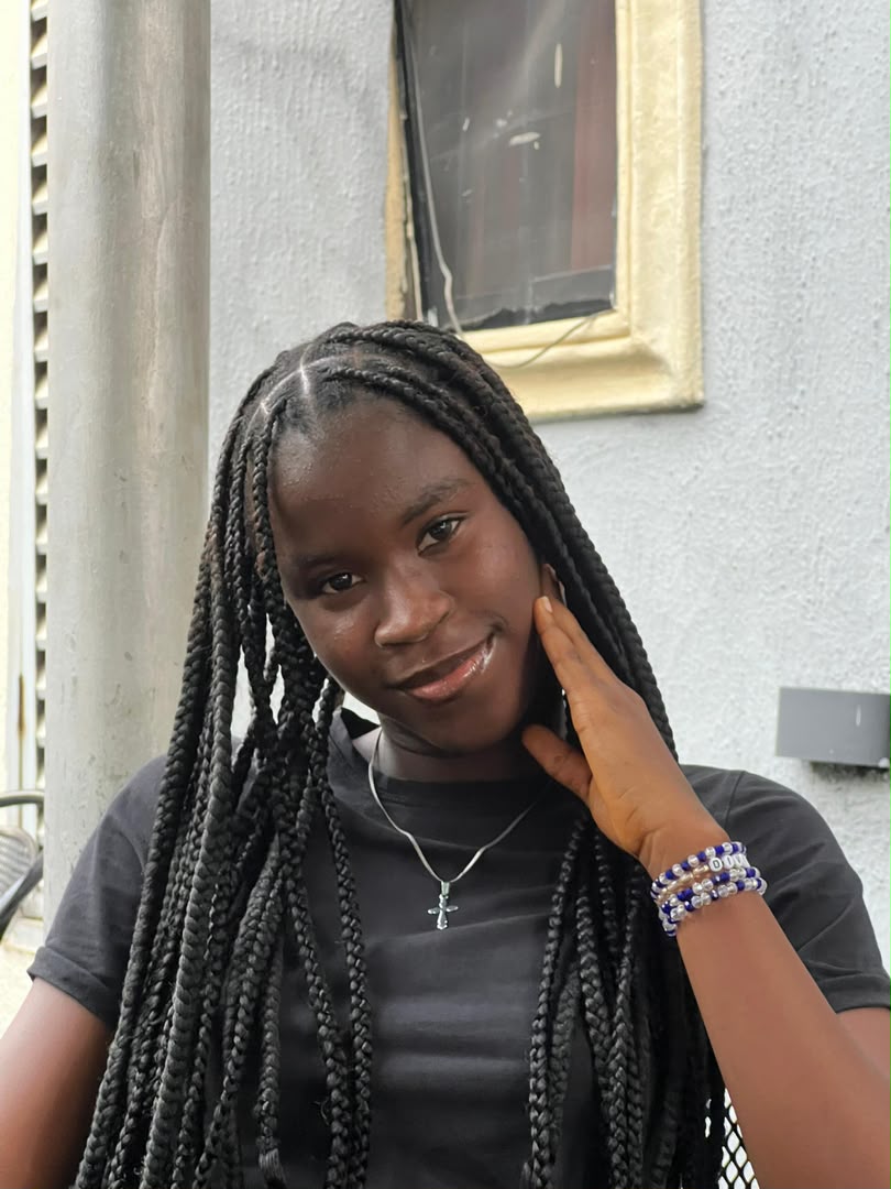 Divine in a black t-shirt with long knotless braids, one hand resting against her cheek.